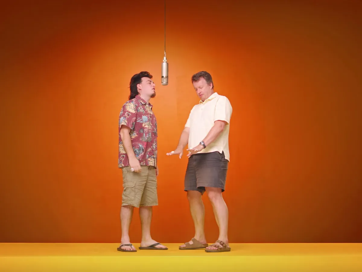 Wide generated frame with Palmer on the left in his red Hawaiian shirt, khaki cargo shorts and flip flops, and Paul on the right in a pale polo, gray shorts and brown sandals