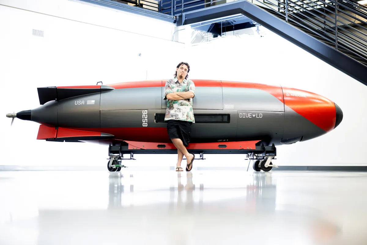 Full body photo of Palmer Luckey in a pale Hawaiian shirt, black shorts and flip flops beside an underwater vehicle