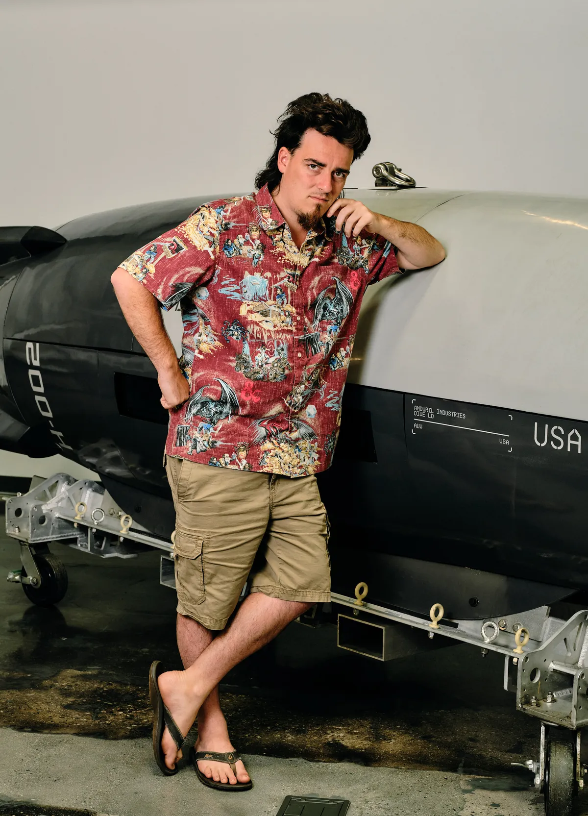 Full body photo of Palmer Luckey in a red Hawaiian shirt, khaki cargo shorts and dark thong flip flops, with both feet visible
