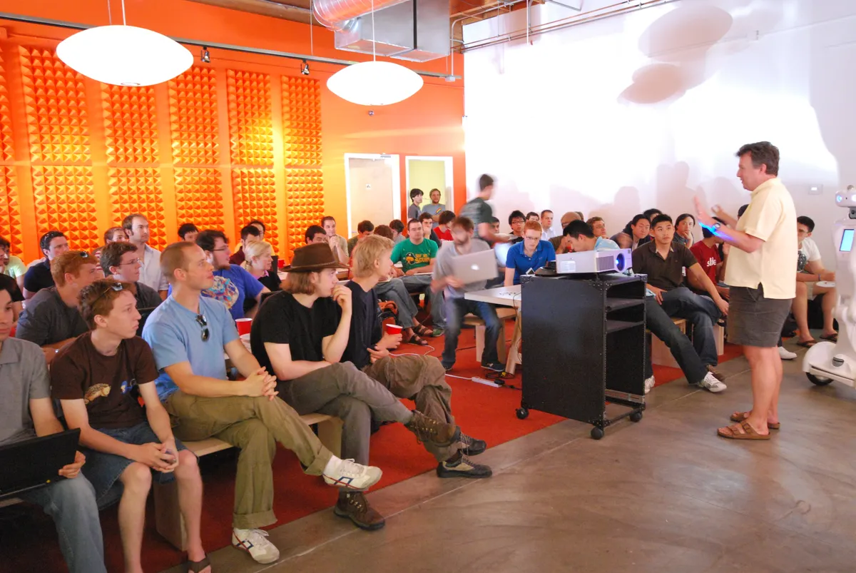 Paul Graham standing at the far right of a YC room in a pale polo, gray shorts and brown sandals